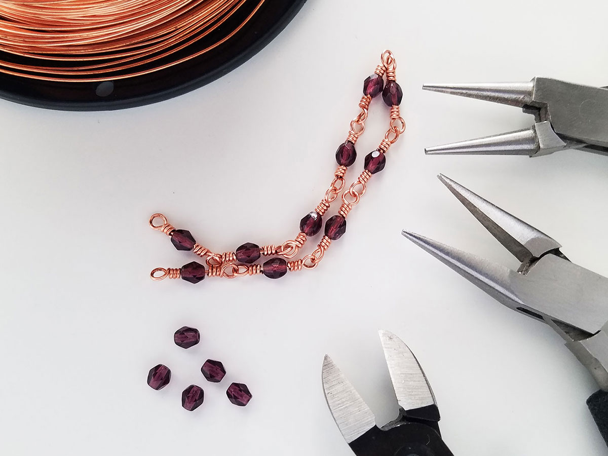 An image of a segment of beaded chain, loose beads, tools and wire used to make the symmetrical wrapped loop connectors demonstrated in this tutorial